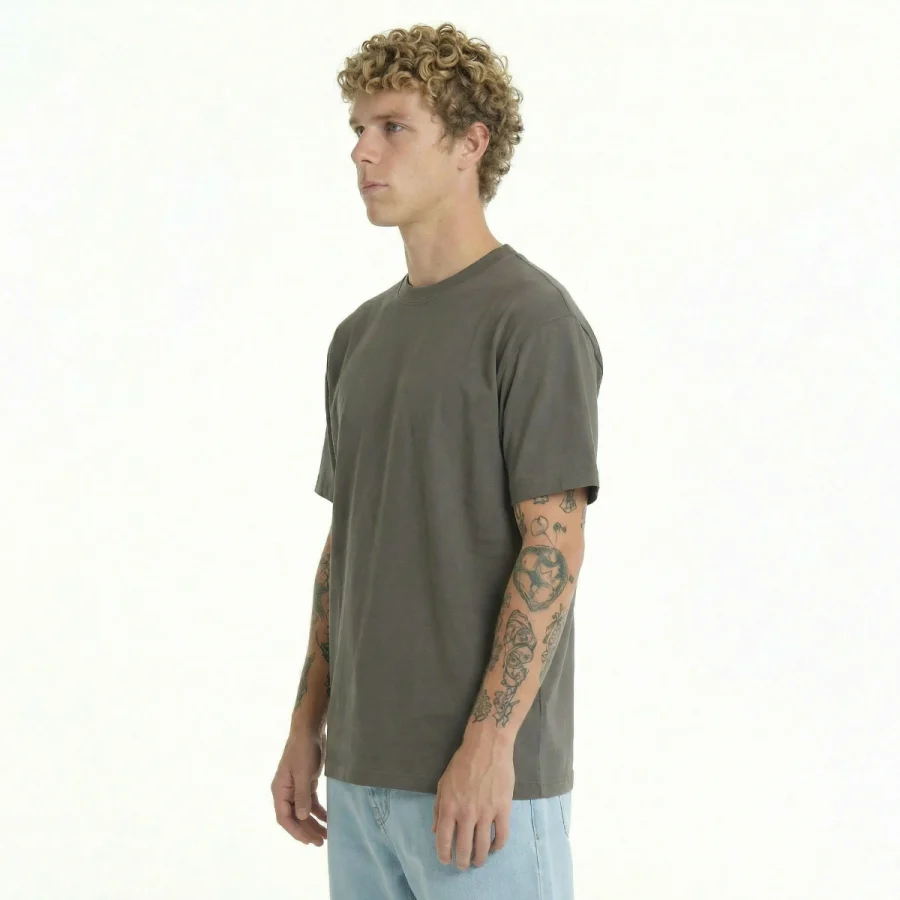 Homem com cabelo encaracolado e tatuagens, vestindo T-Shirt Over Relaxed Fit cinza escura e jeans claros.