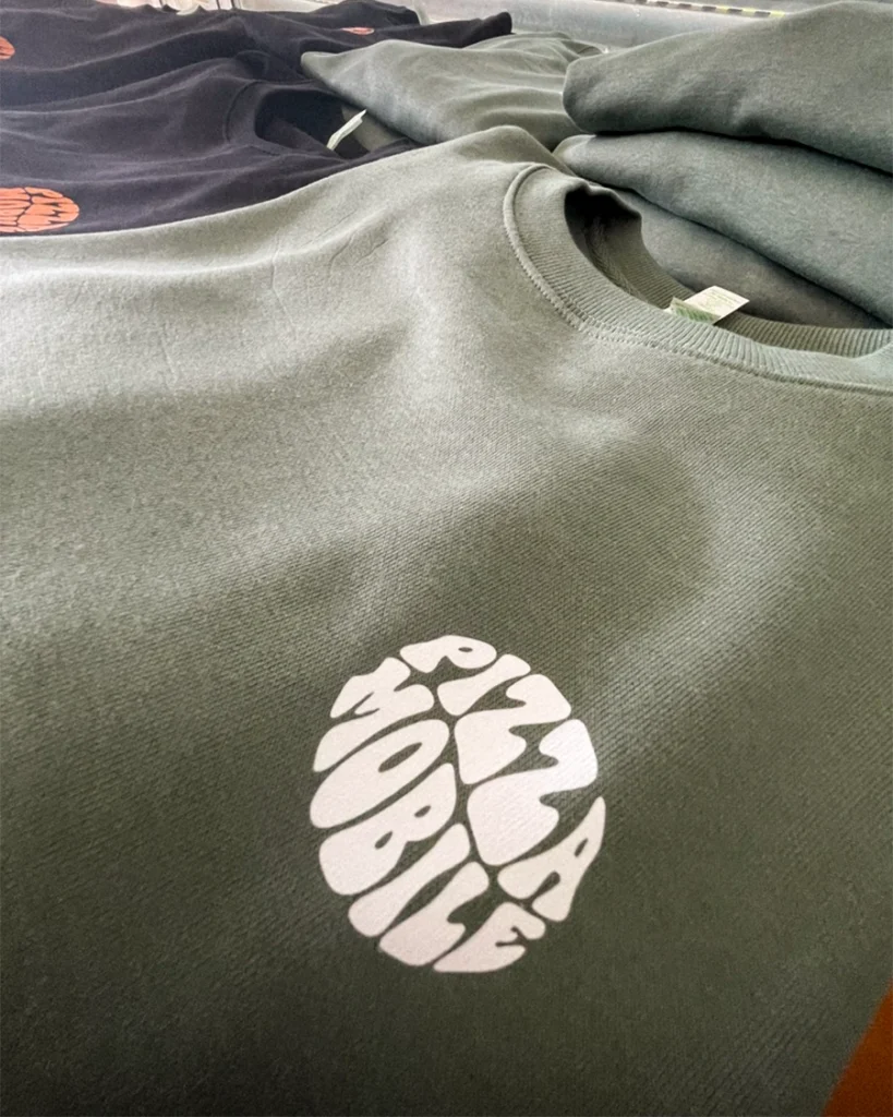Camisola verde com logótipo "Pizza Mobile" em branco, empilhada com outras roupas.