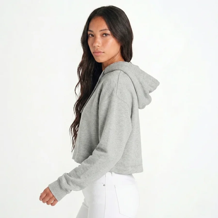 Mulher com cabelo castanho, de costas viradas para a direita e virada para a câmara, vestindo hoodie cropped cinzento e calças brancas.