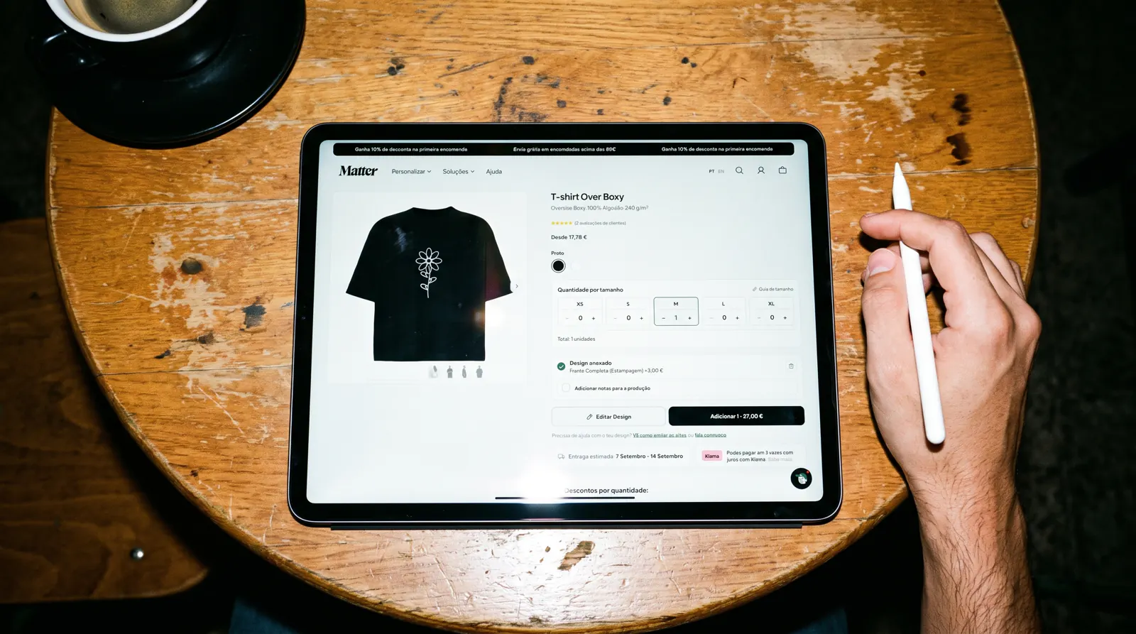 iPad numa mesa de café a mostrar o personalizador do Matter Studio com uma t-shirt oversize preta com uma flor em linha branca, uma unidade no carrinho
